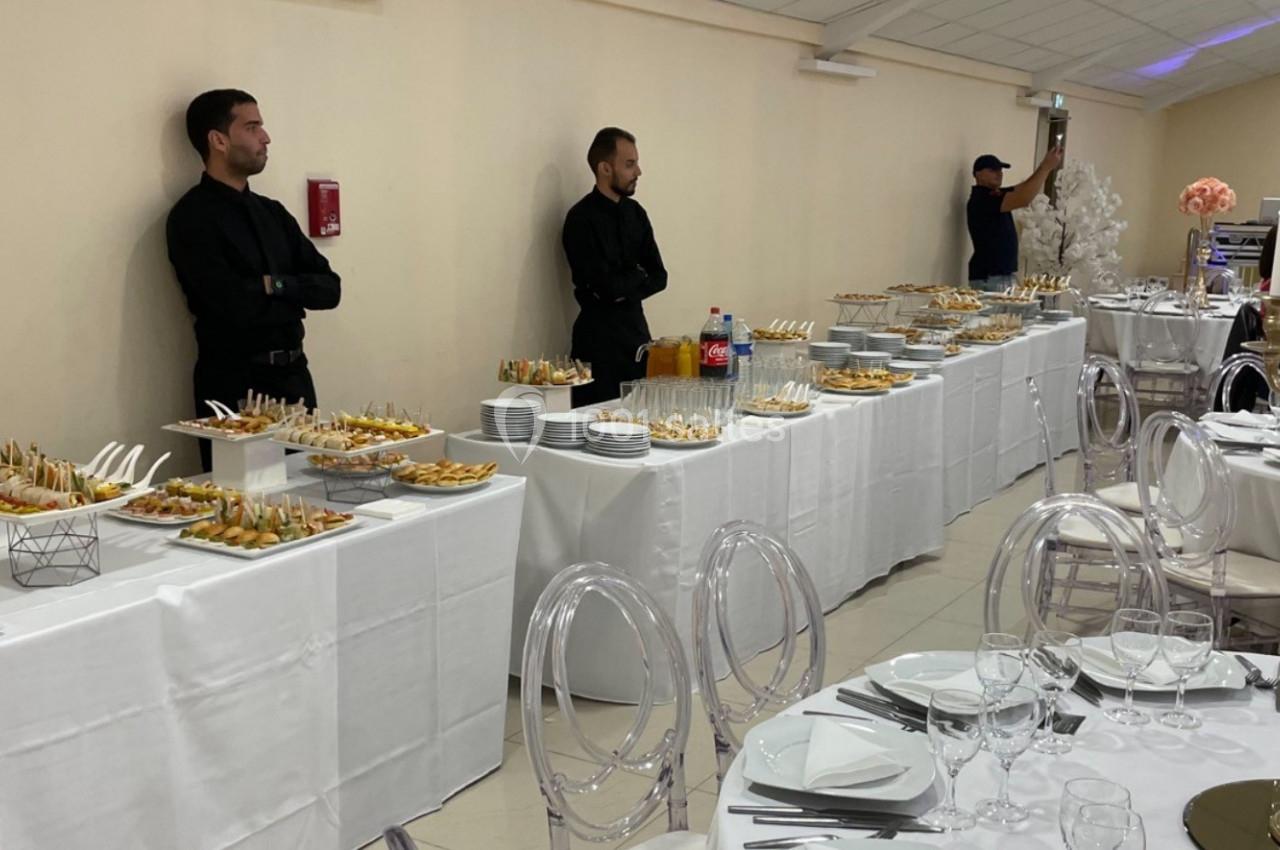 Buffet de réception avec plats variés, serveurs en tenue noire et tables dressées pour les invités.