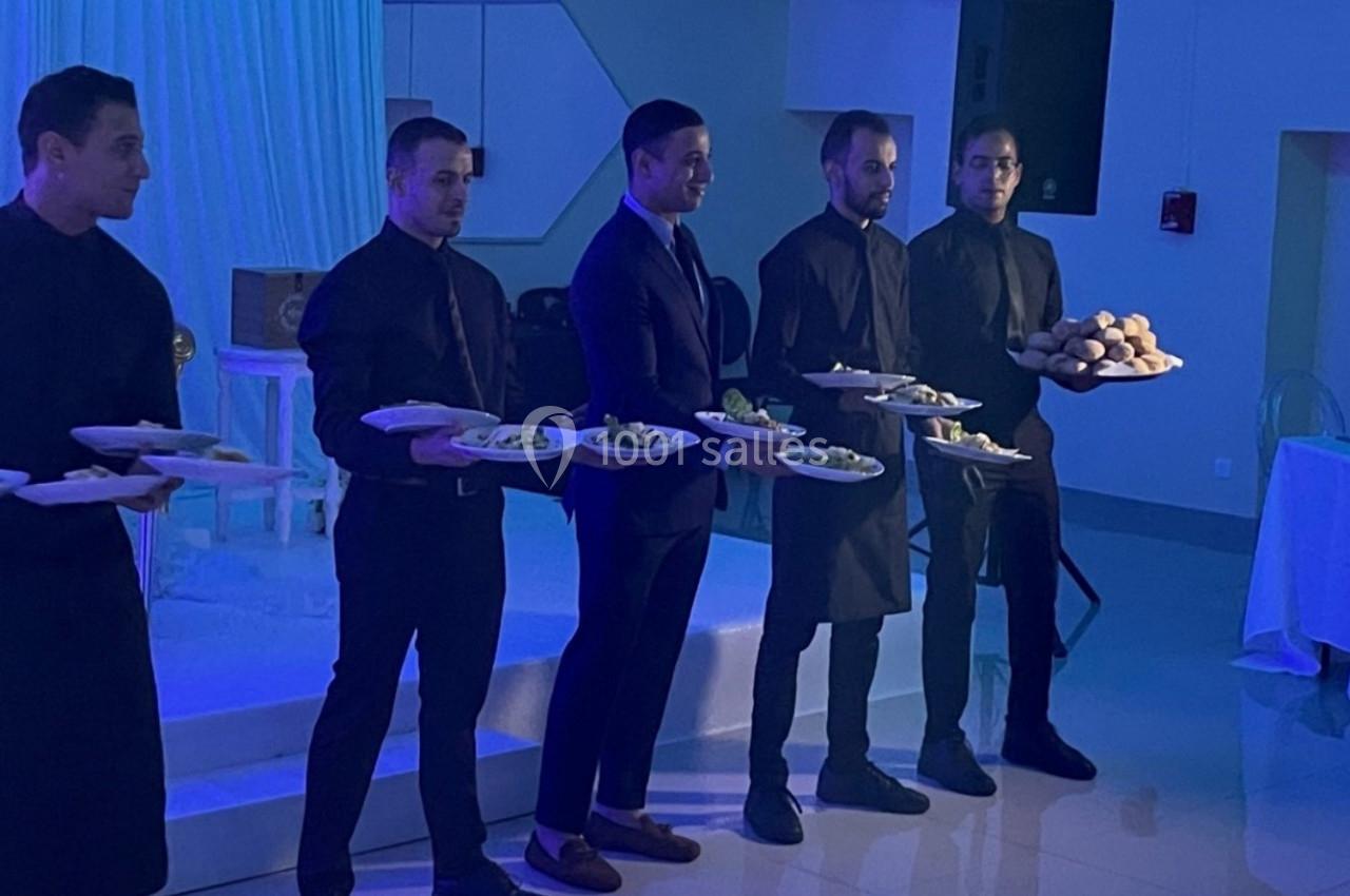 Des serveurs en tenue formelle portant des assiettes de plats dans une salle éclairée par une lumière bleutée.