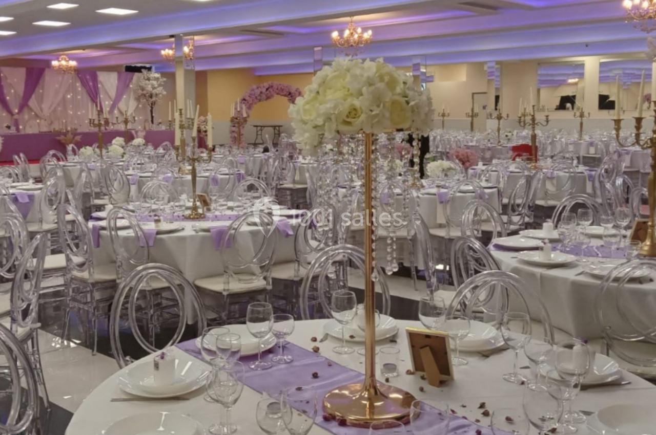Salle de réception décorée avec des tables rondes, nappes blanches, chaises transparentes et centres de table fleuris.