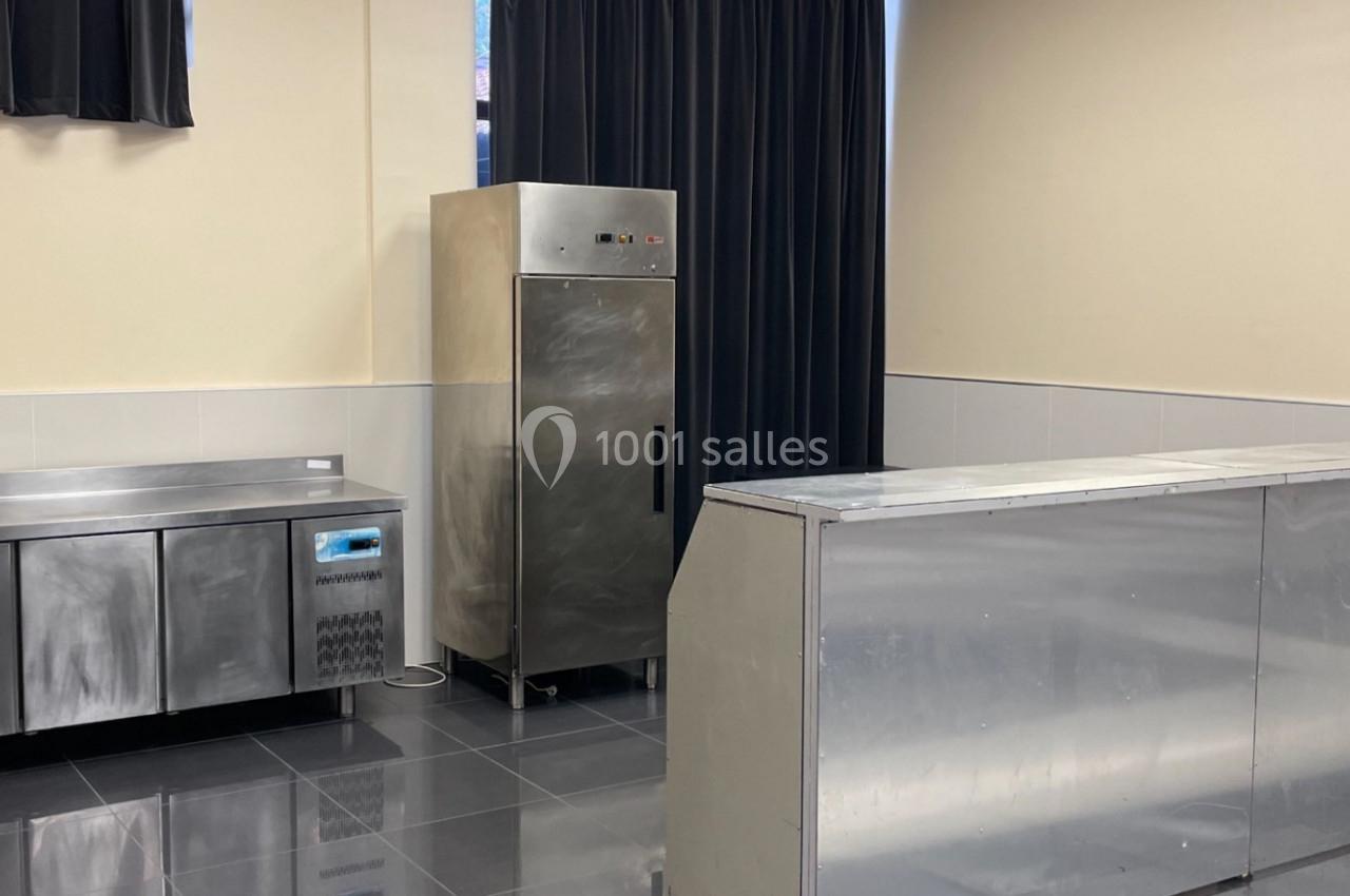 Cuisine professionnelle avec comptoirs en inox, réfrigérateur vertical et carrelage gris foncé.