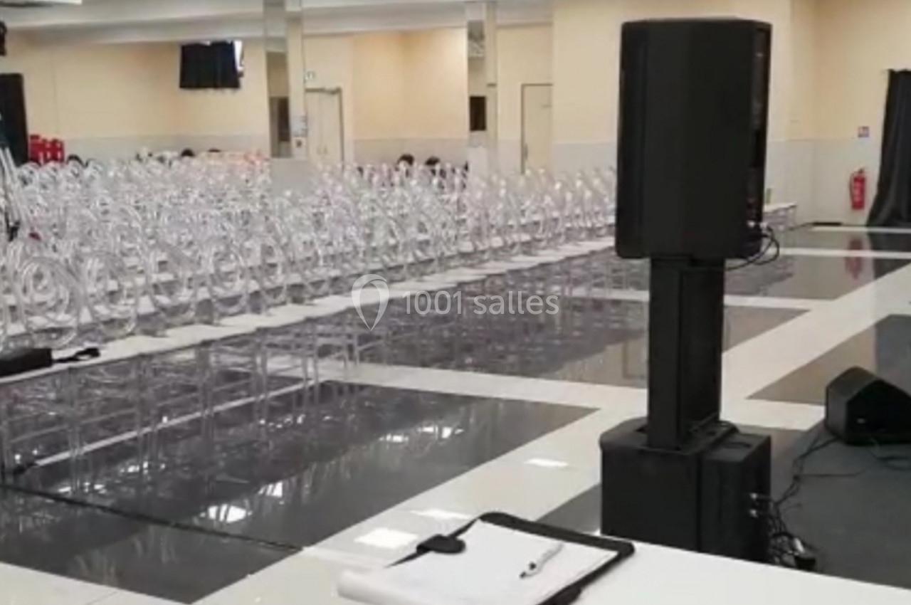 Salle de conférence vide avec des rangées de chaises transparentes, un podium, des enceintes et un sol réfléchissant.