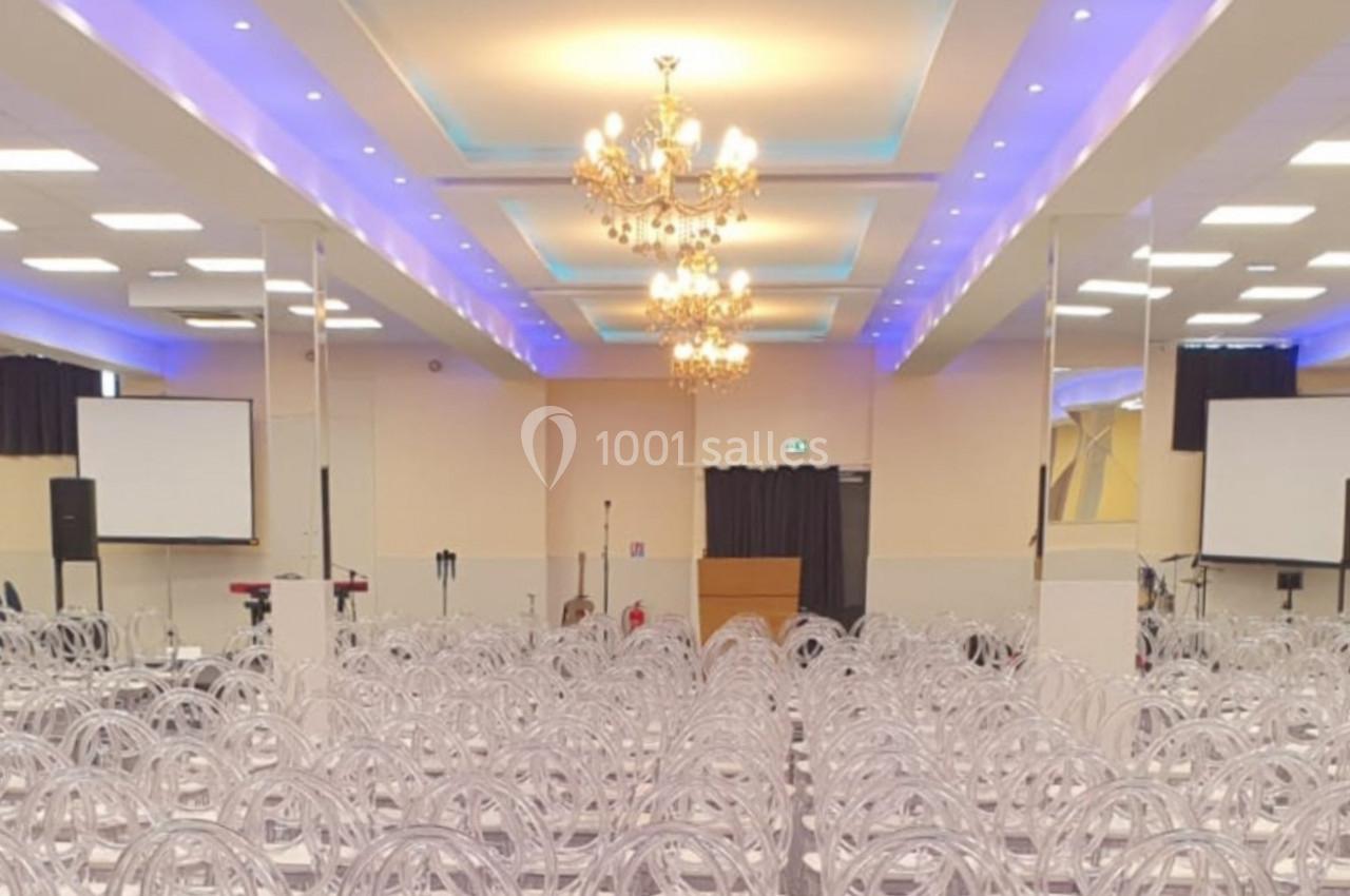 Salle de conférence lumineuse avec rangées de chaises transparentes, écrans de projection et lustres au plafond.