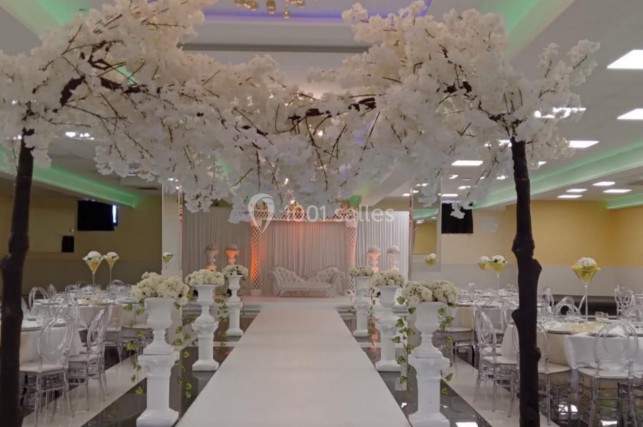 Allée centrale décorée de fleurs blanches menant à une scène dans une salle de réception élégamment éclairée.