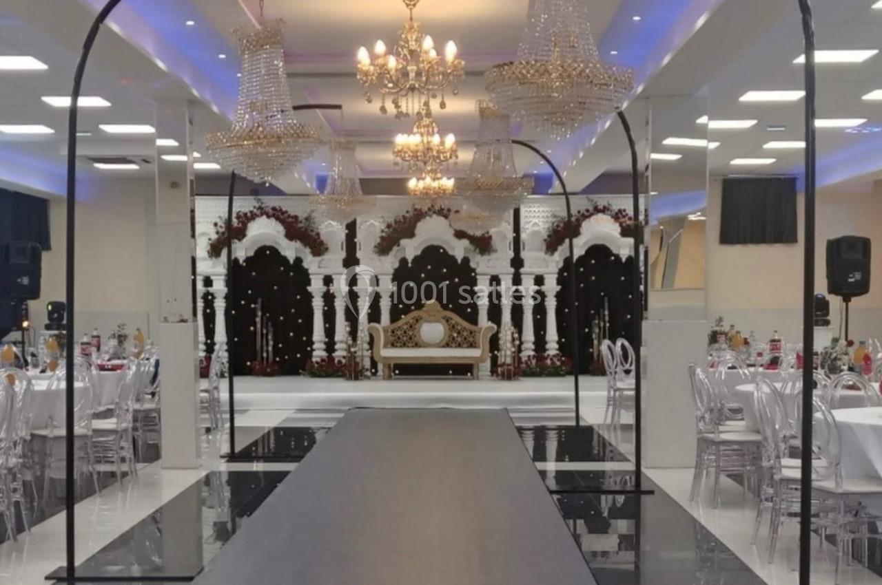 Salle de réception décorée avec des lustres, une scène ornée d'arcs blancs et un canapé, tables dressées autour.