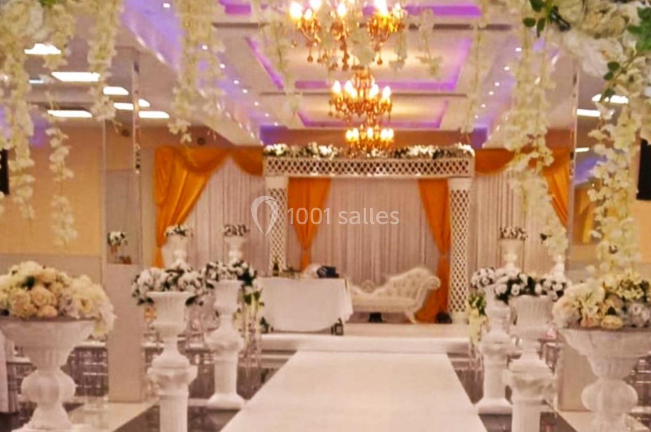 Allée décorée de fleurs blanches menant à une arche et une scène ornée pour une cérémonie dans une salle de réception.