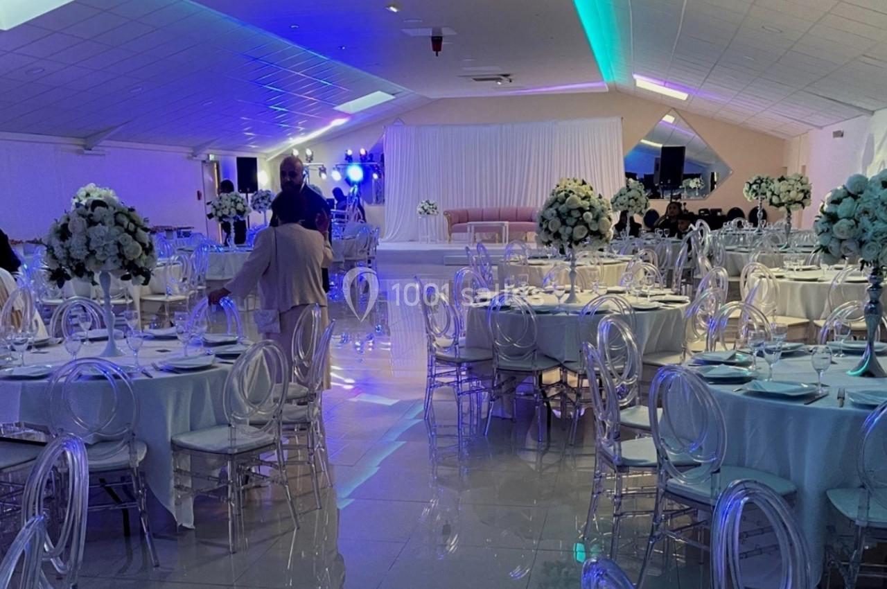 Salle de réception décorée avec des tables rondes, nappes blanches, chaises transparentes et arrangements floraux.