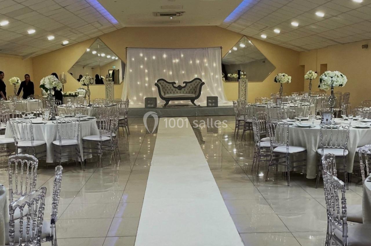 Salle de réception élégante avec tables rondes dressées, allée centrale blanche et canapé décoratif sur une estrade.