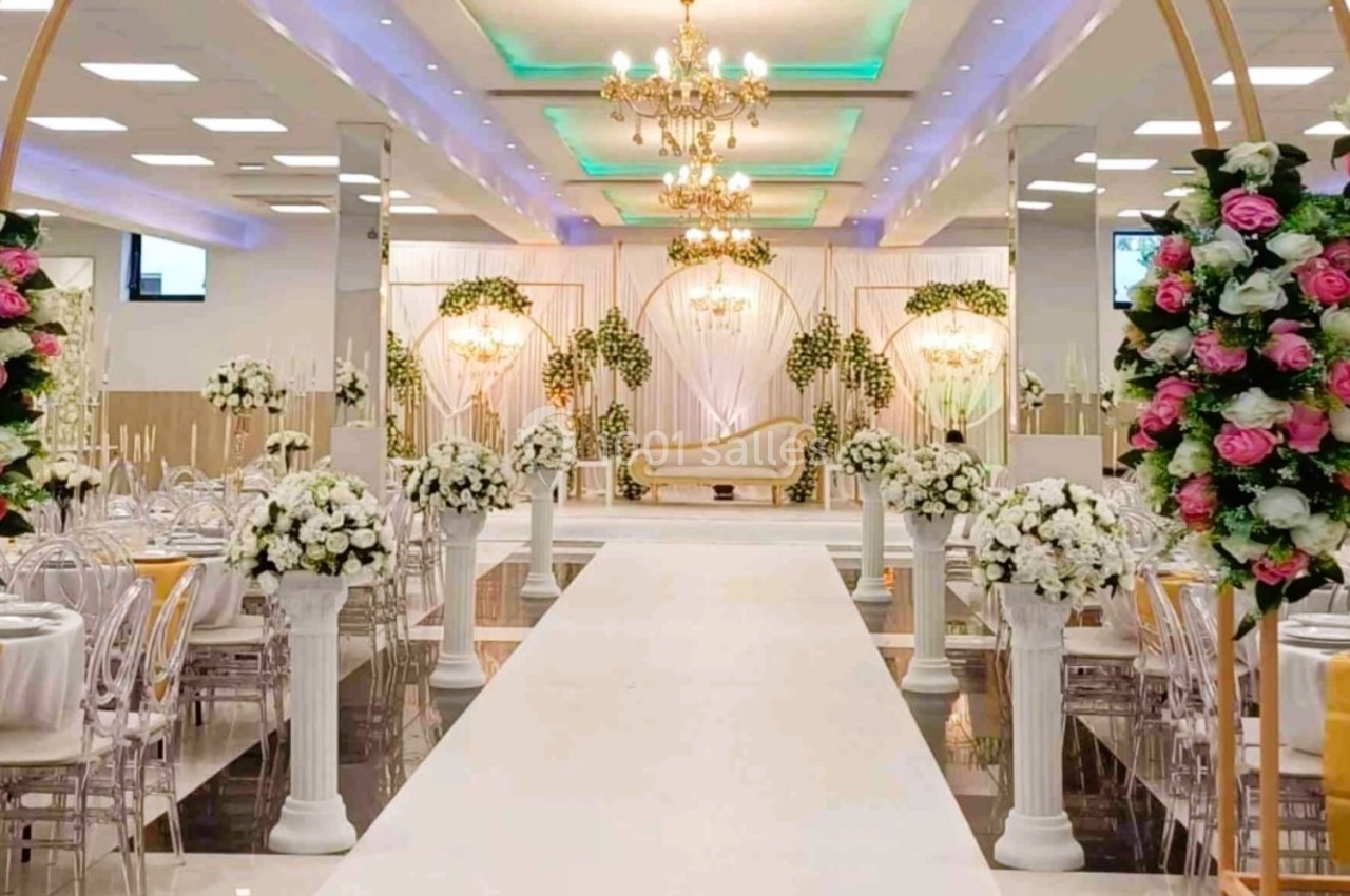 Salle de réception décorée avec des arches florales, des chandeliers et une allée centrale menant à une estrade ornée.