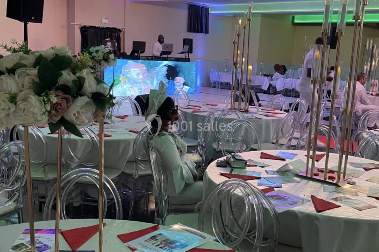 Salle de réception décorée avec des tables rondes, nappes blanches, fleurs et bougies, avec des invités assis.