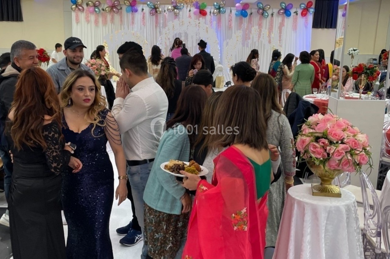 Groupe de personnes discutant et mangeant dans une salle décorée pour un événement festif avec des tables et des fleurs.