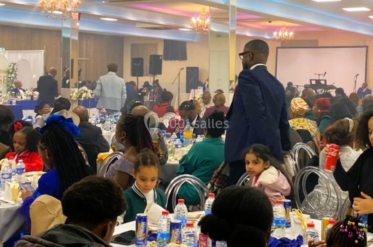 Salle de réception bondée avec des invités assis à des tables décorées, participant à un événement festif.