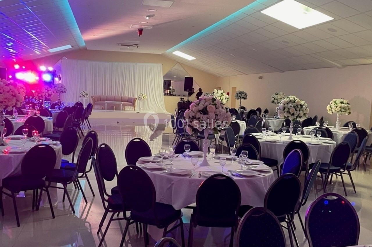 Salle de réception décorée avec des tables rondes dressées, des chaises noires et des arrangements floraux blancs.
