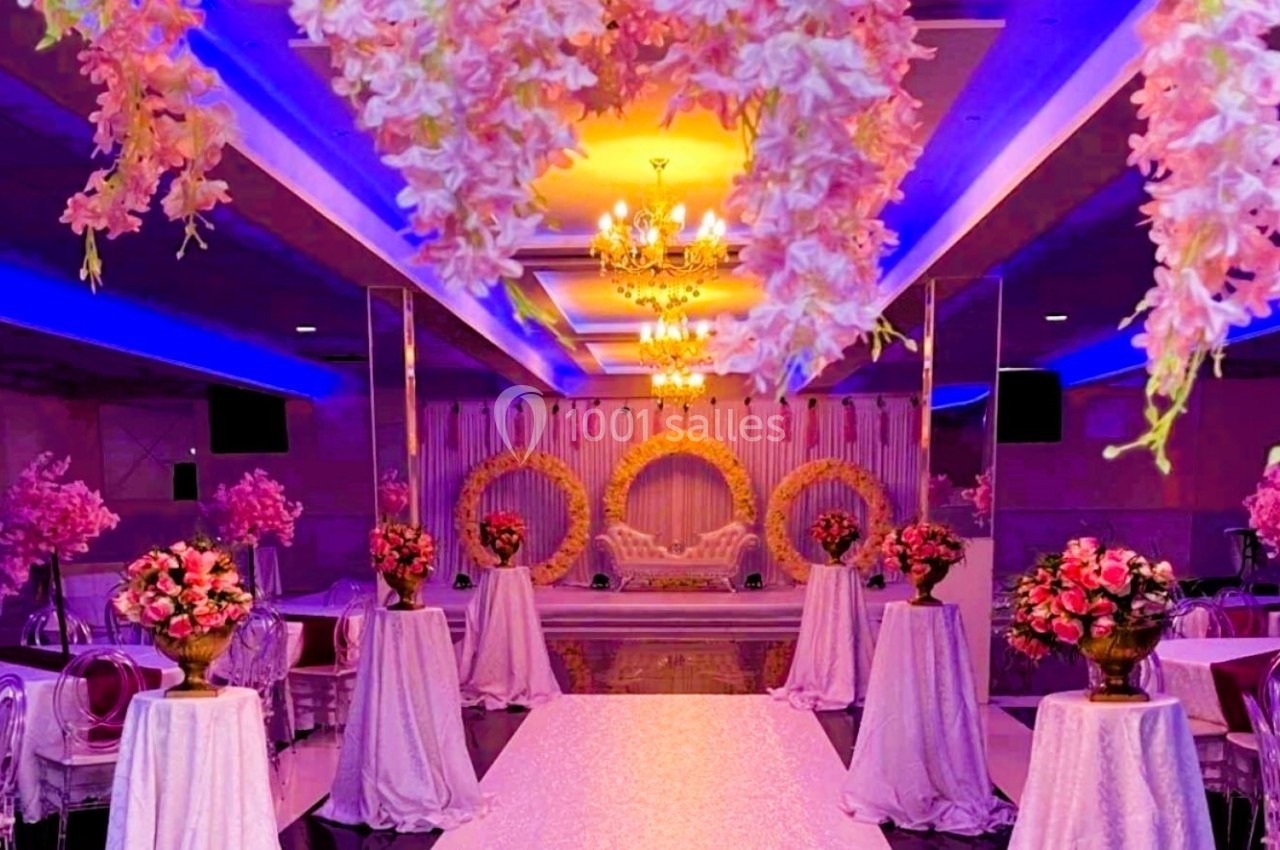 Salle de réception décorée avec des fleurs suspendues, des tables ornées de bouquets et un éclairage coloré.
