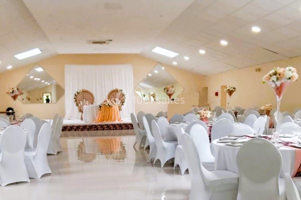 Salle de réception décorée avec des tables rondes, nappes blanches, centres de table fleuris et une scène ornée.