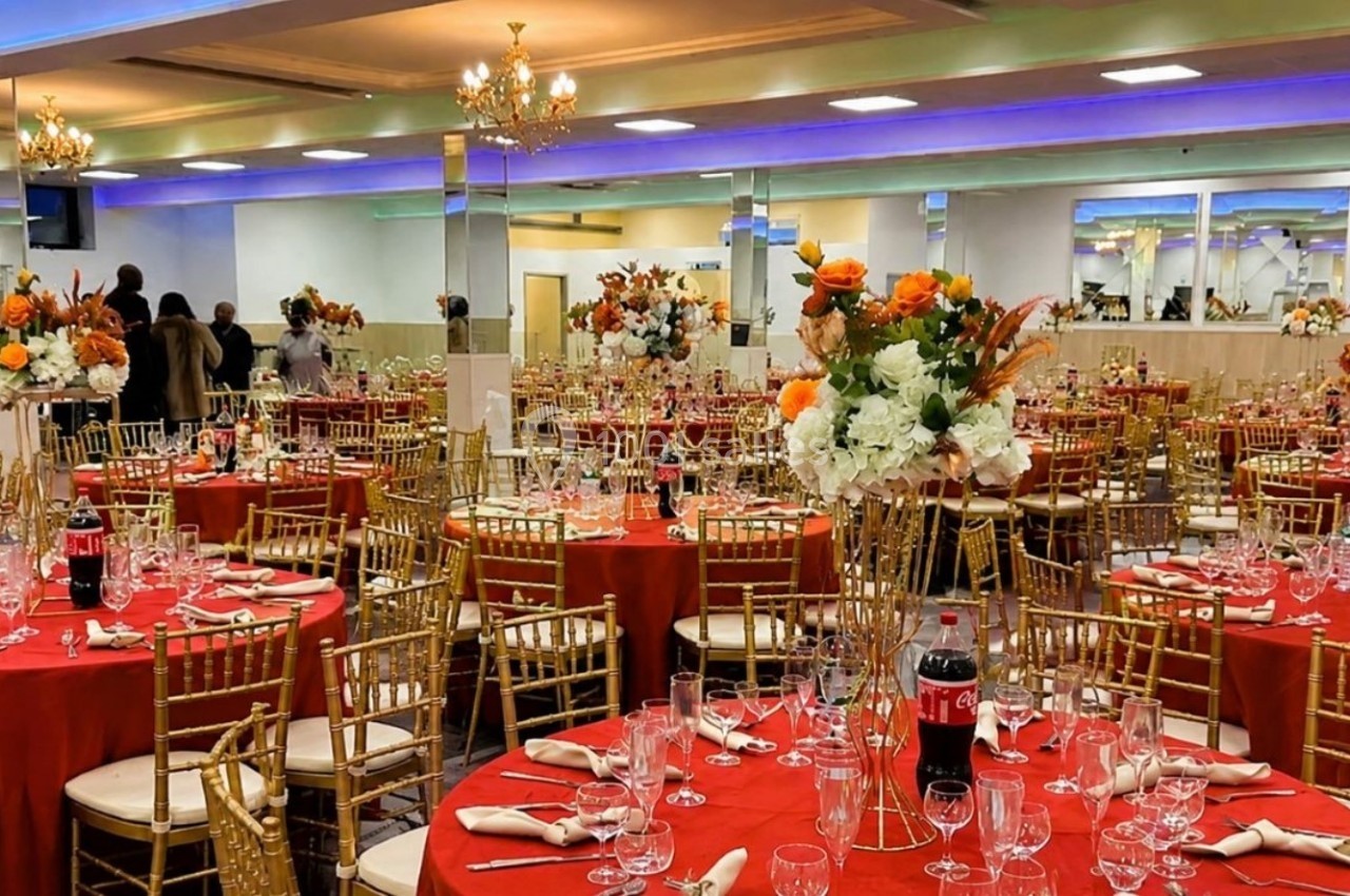 Salle de réception décorée avec des tables rondes, nappes rouges, chaises dorées et centres de table fleuris.