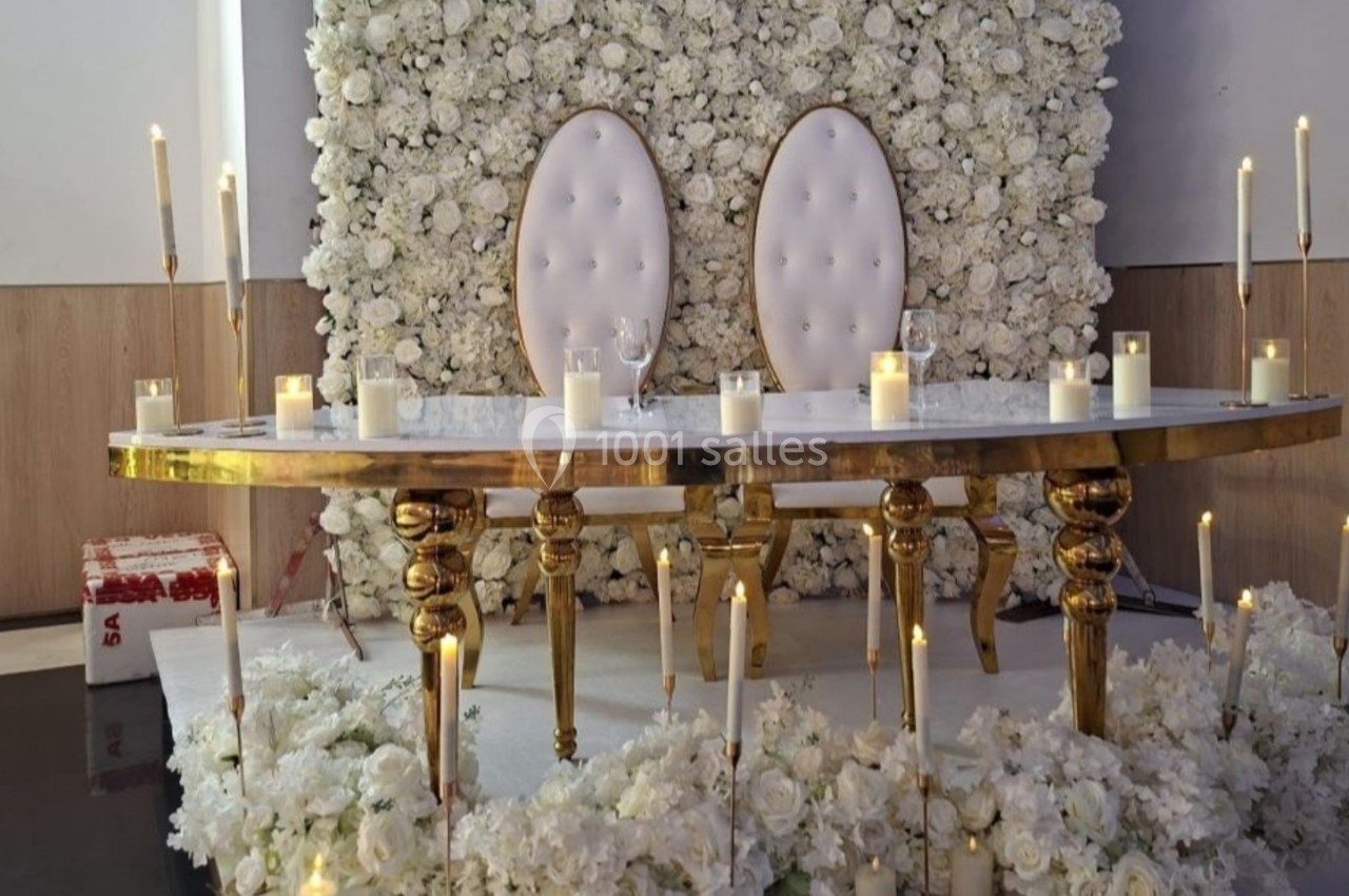 Table décorée avec des bougies et des fleurs blanches devant un mur floral, deux chaises blanches en arrière-plan.
