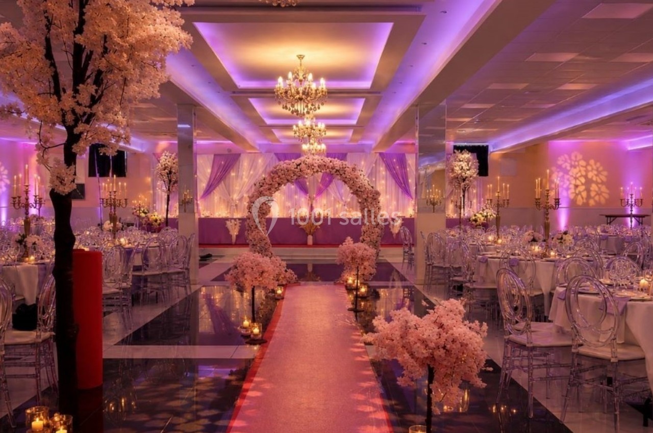 Salle de réception élégante avec éclairage violet, chandeliers, arche florale et allée décorée de fleurs roses.