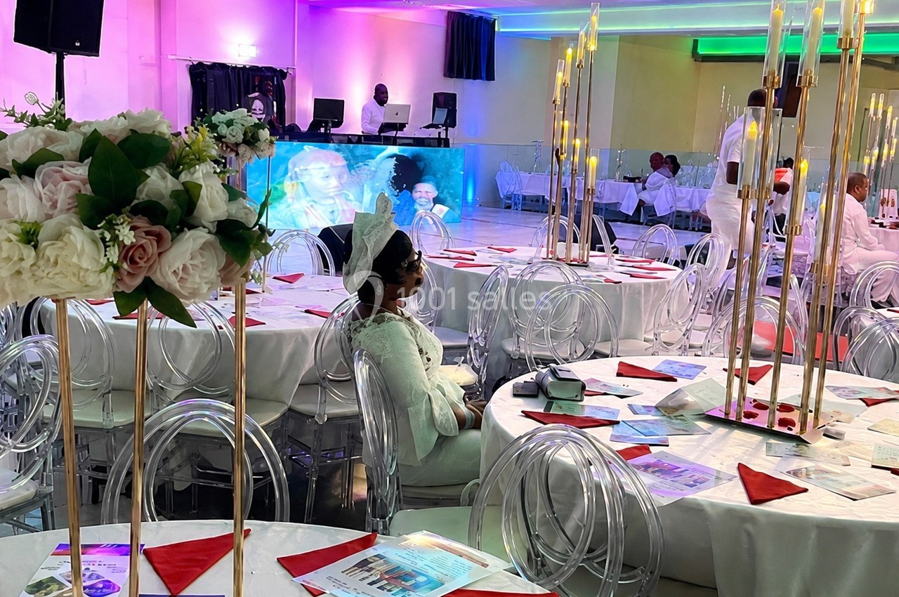Salle de réception décorée avec des tables rondes, nappes blanches, bouquets de fleurs et chandeliers, invités assis.