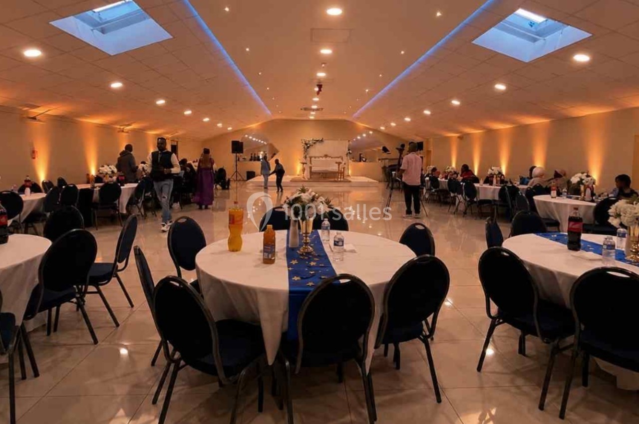 Salle de réception éclairée, décorée avec des tables rondes, nappes blanches et centres de table fleuris.