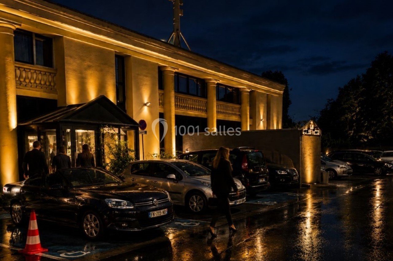 Façade d'un bâtiment éclairé de nuit avec des voitures garées sur un parking mouillé par la pluie.