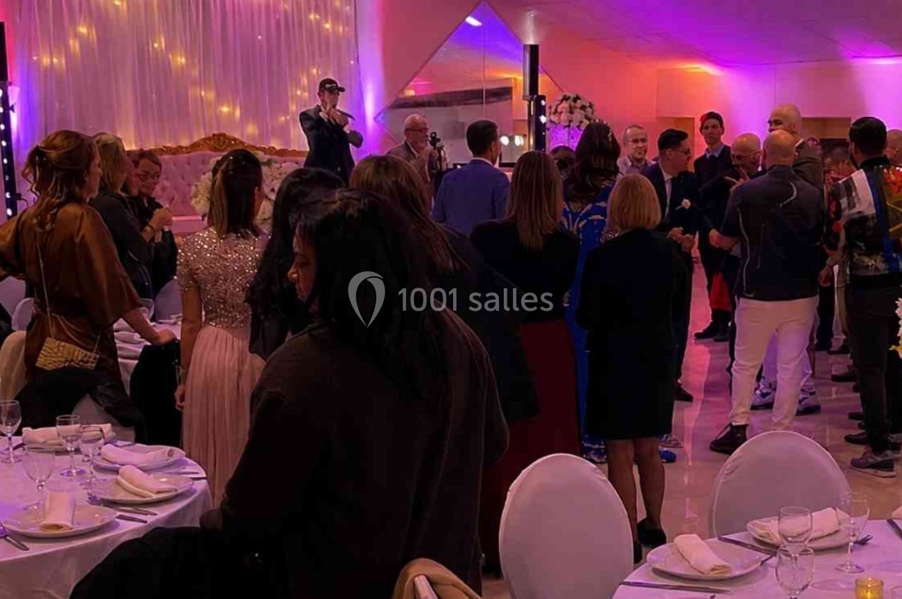 Des invités debout dans une salle de réception décorée avec des fleurs et des lumières colorées, écoutant un discours.