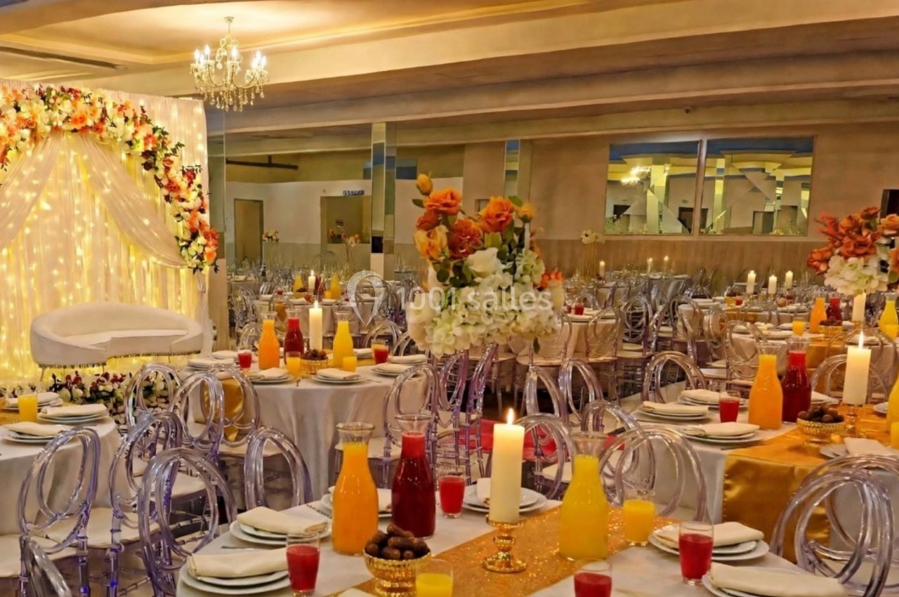 Salle de réception décorée avec des tables dressées, des chaises transparentes et un fond floral illuminé.