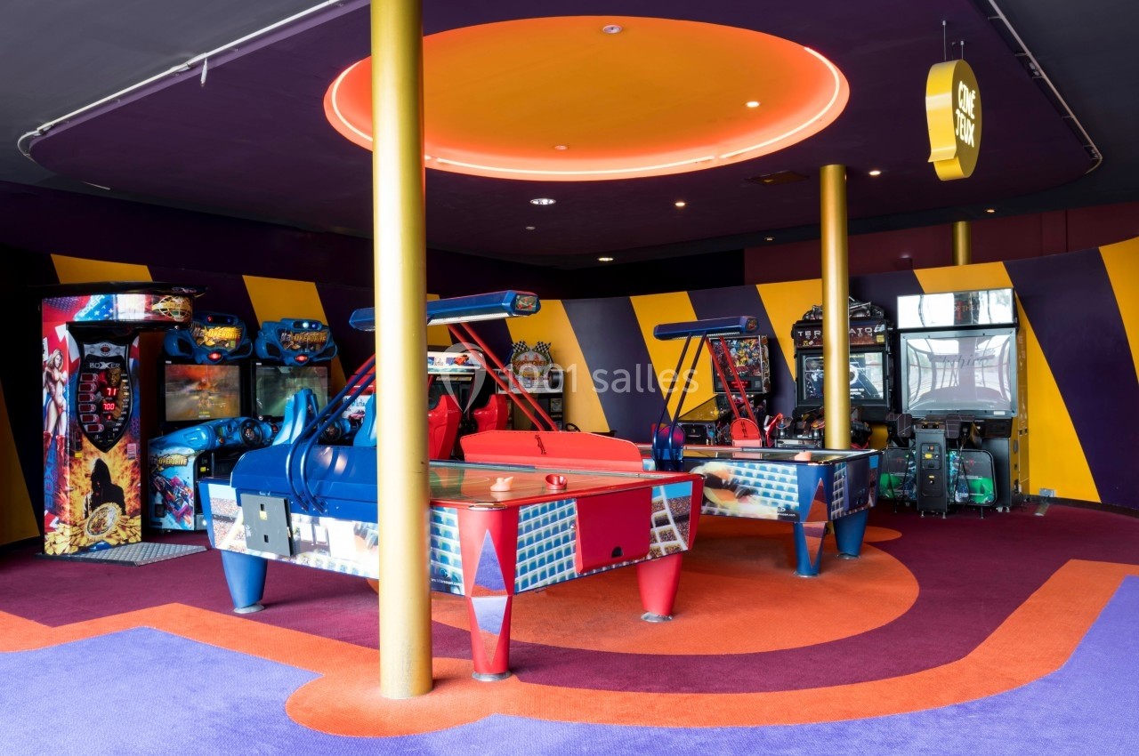 Salle d'arcade colorée avec des machines de jeux, des tables de hockey sur air et un éclairage néon au plafond.