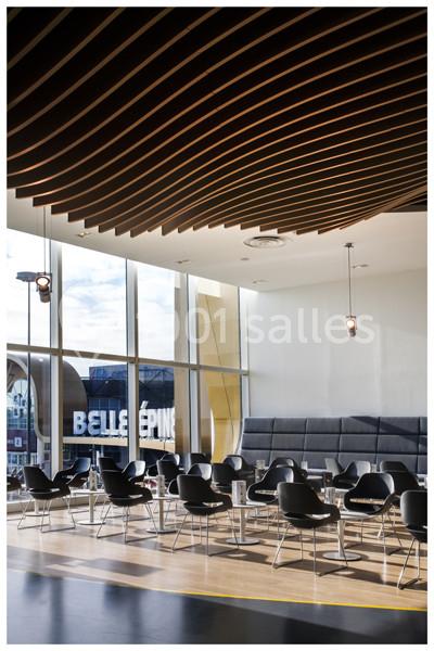 Salle moderne avec des chaises noires, tables basses et plafond ondulé en bois, éclairée par de grandes fenêtres.