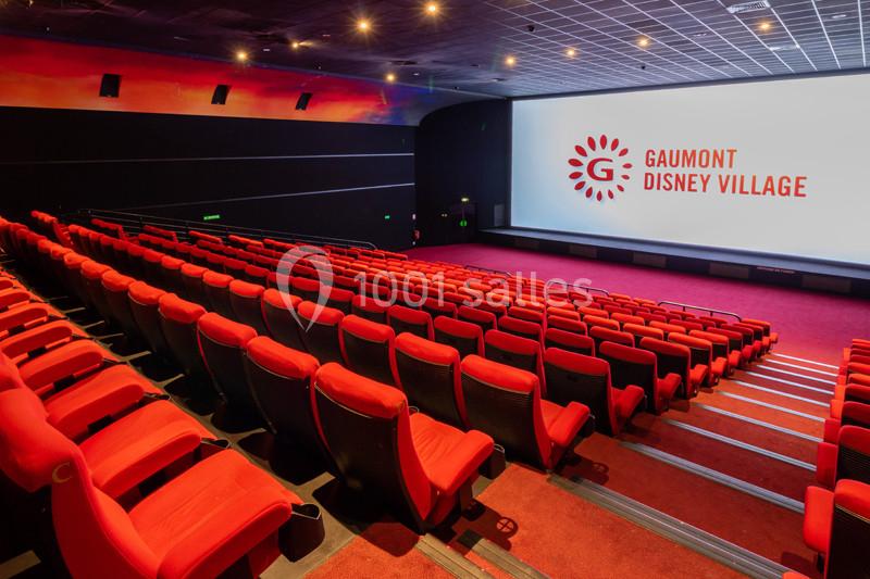 Salle de cinéma vide avec rangées de fauteuils rouges, grand écran allumé affichant un logo et un plafond éclairé.