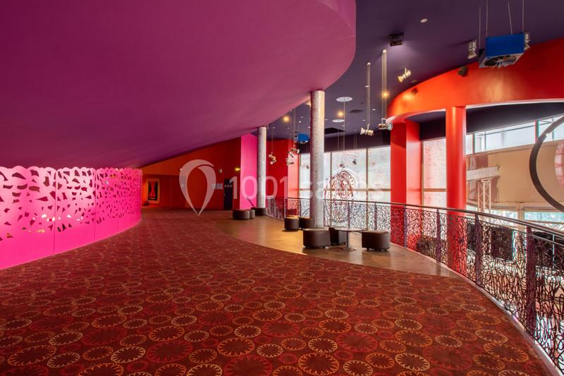 Couloir spacieux avec moquette rouge à motifs, murs colorés et balustrade métallique, éclairé par des fenêtres.