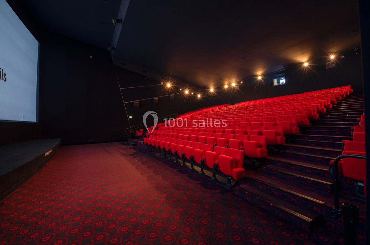 Salle de cinéma vide avec des rangées de sièges rouges et un écran blanc allumé à gauche.