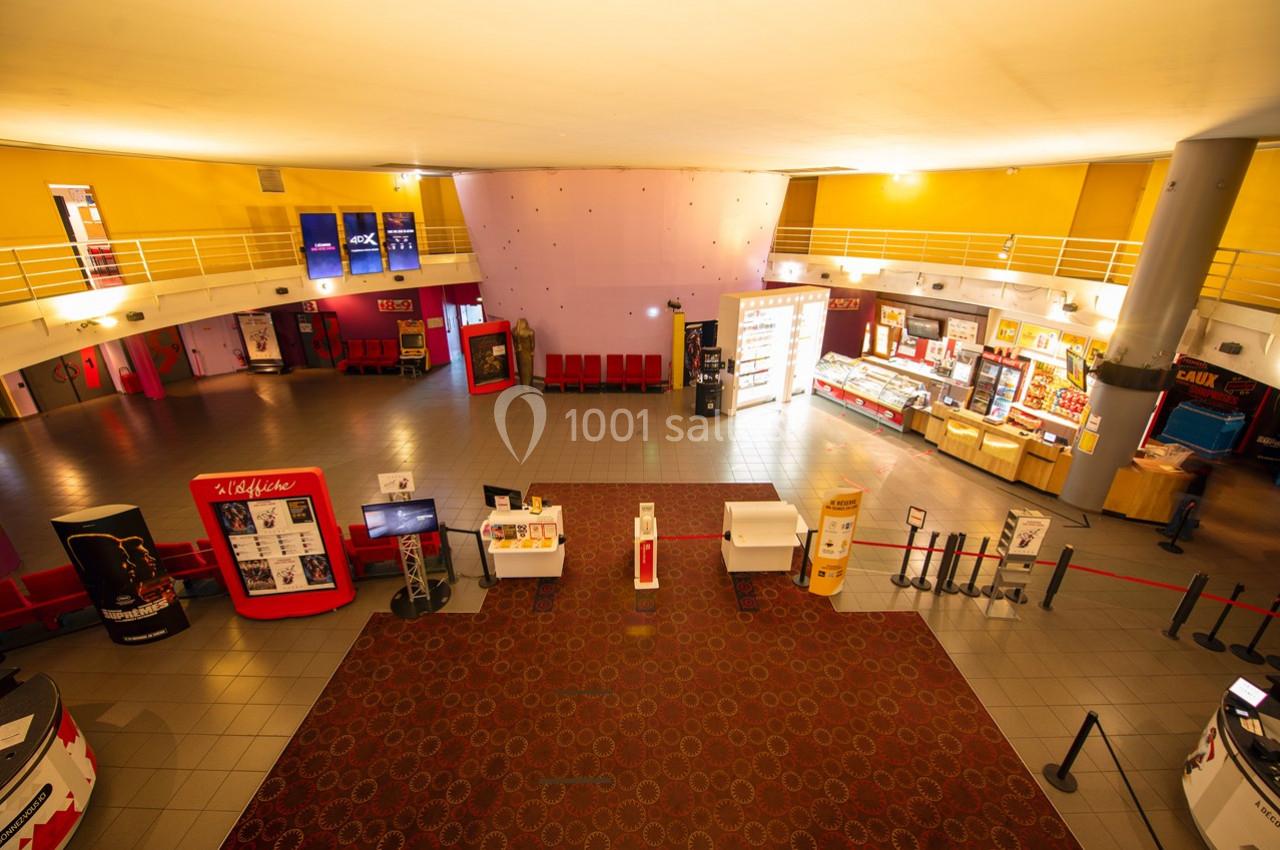 Hall d'entrée d'un cinéma avec comptoirs de confiserie, bornes interactives et rangées de sièges rouges.
