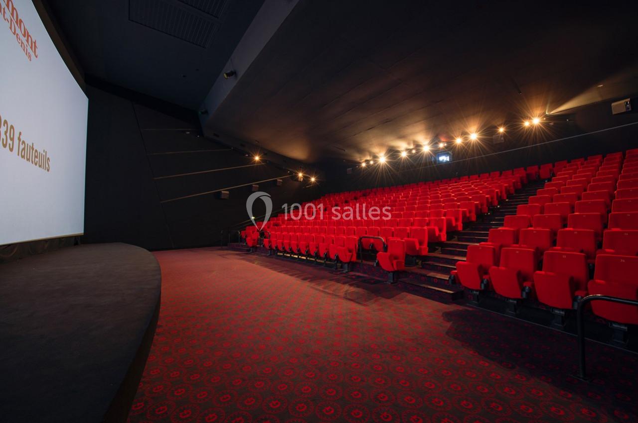 Salle de cinéma vide avec des rangées de fauteuils rouges et un grand écran blanc sur la gauche.