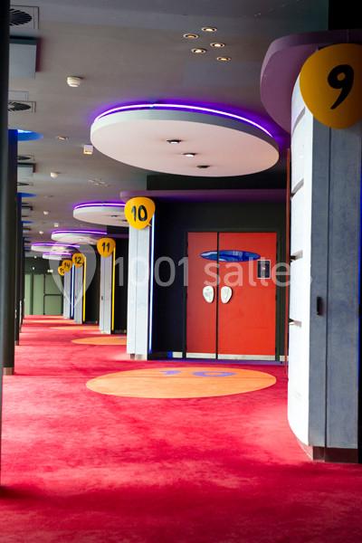 Couloir d'un cinéma avec moquette rouge, portes numérotées et éclairage violet au plafond.