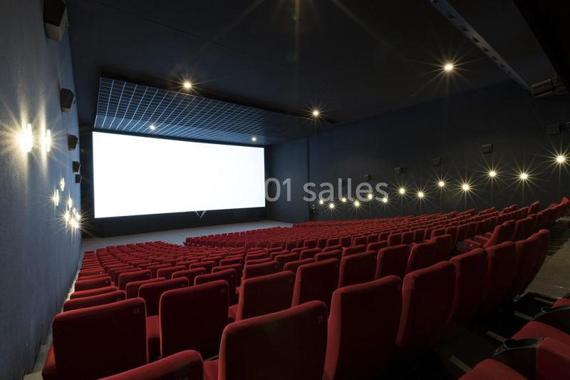 Salle de cinéma vide avec rangées de sièges rouges et un grand écran blanc allumé.