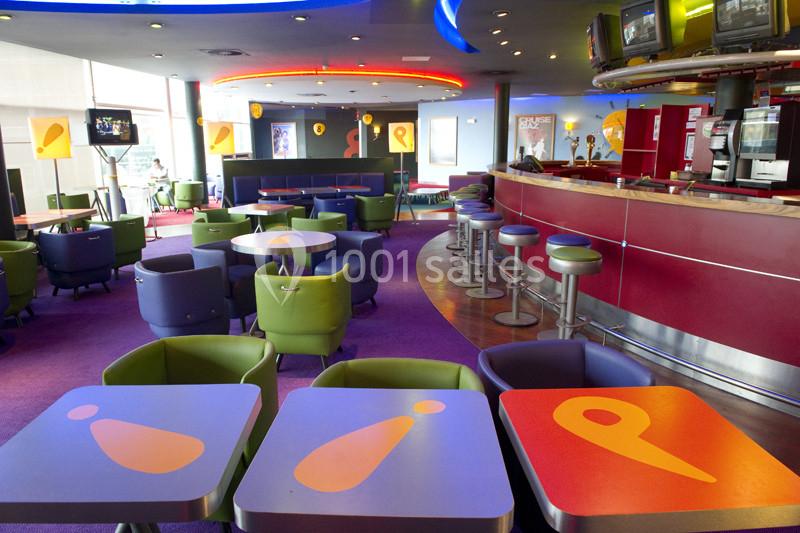 Salle colorée avec tables décorées, fauteuils, bar rouge et écrans suspendus dans un espace moderne et lumineux.