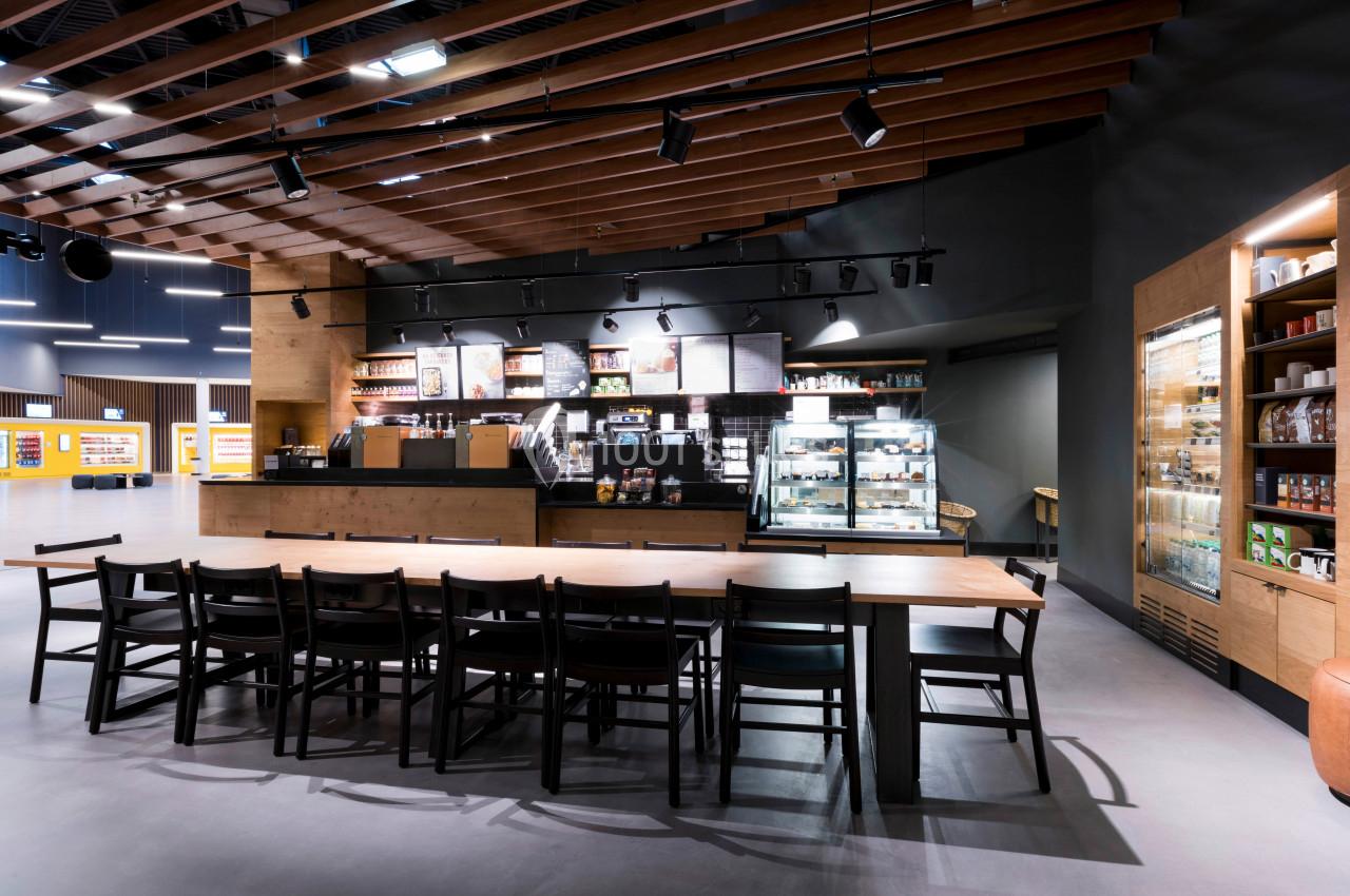 Intérieur d'un café moderne avec un comptoir, des étagères remplies de produits et une grande table en bois avec des chaises.