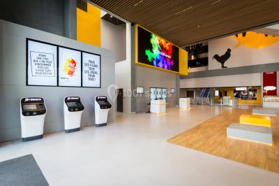 Hall moderne avec bancs, écrans numériques, visiteurs et décorations colorées, incluant une silhouette de coq sur un mur.