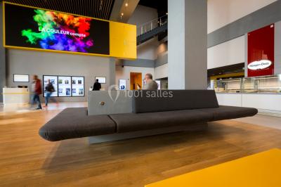 Hall moderne avec bancs, écrans numériques, visiteurs et décorations colorées, incluant une silhouette de coq sur un mur.