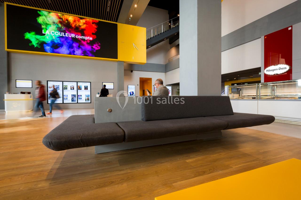 Un hall moderne avec un canapé gris, un écran affichant des couleurs vives et des visiteurs en arrière-plan.