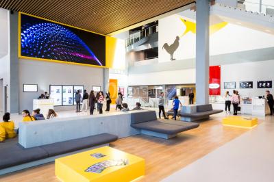 Hall moderne avec bancs, écrans numériques, visiteurs et décorations colorées, incluant une silhouette de coq sur un mur.