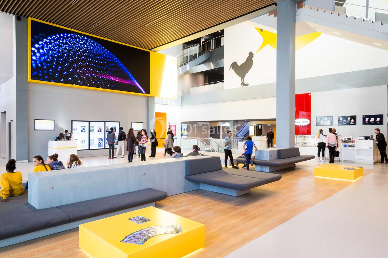 Hall moderne avec bancs, écrans numériques, visiteurs et décorations colorées, incluant une silhouette de coq sur un mur.