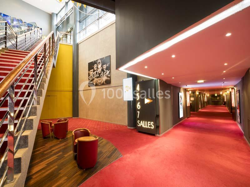 Hall de cinéma avec escalier, fauteuils rouges, affiches murales et couloir menant aux salles numérotées.