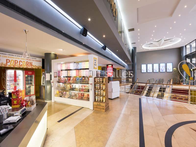 Vue d'un hall de cinéma moderne avec un comptoir de confiseries, des affiches lumineuses et un escalier roulant.