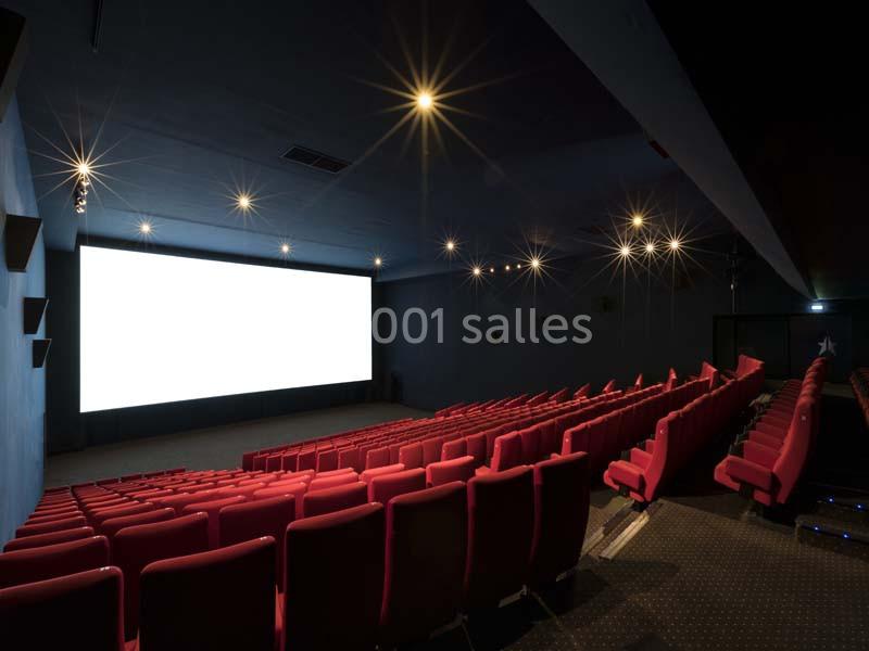 Salle de cinéma vide avec rangées de sièges rouges et un grand écran blanc allumé.