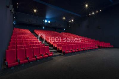 Salle de cinéma vide avec des rangées de sièges rouges et un écran allumé au fond.