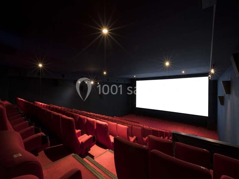 Salle de cinéma vide avec des rangées de sièges rouges et un écran blanc allumé au fond.