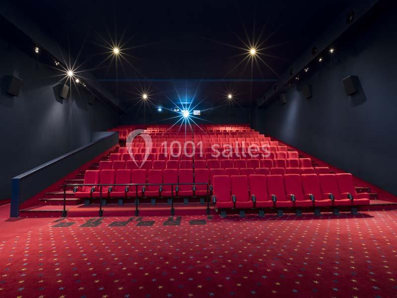 Salle de cinéma vide avec des rangées de sièges rouges et un écran allumé au fond.