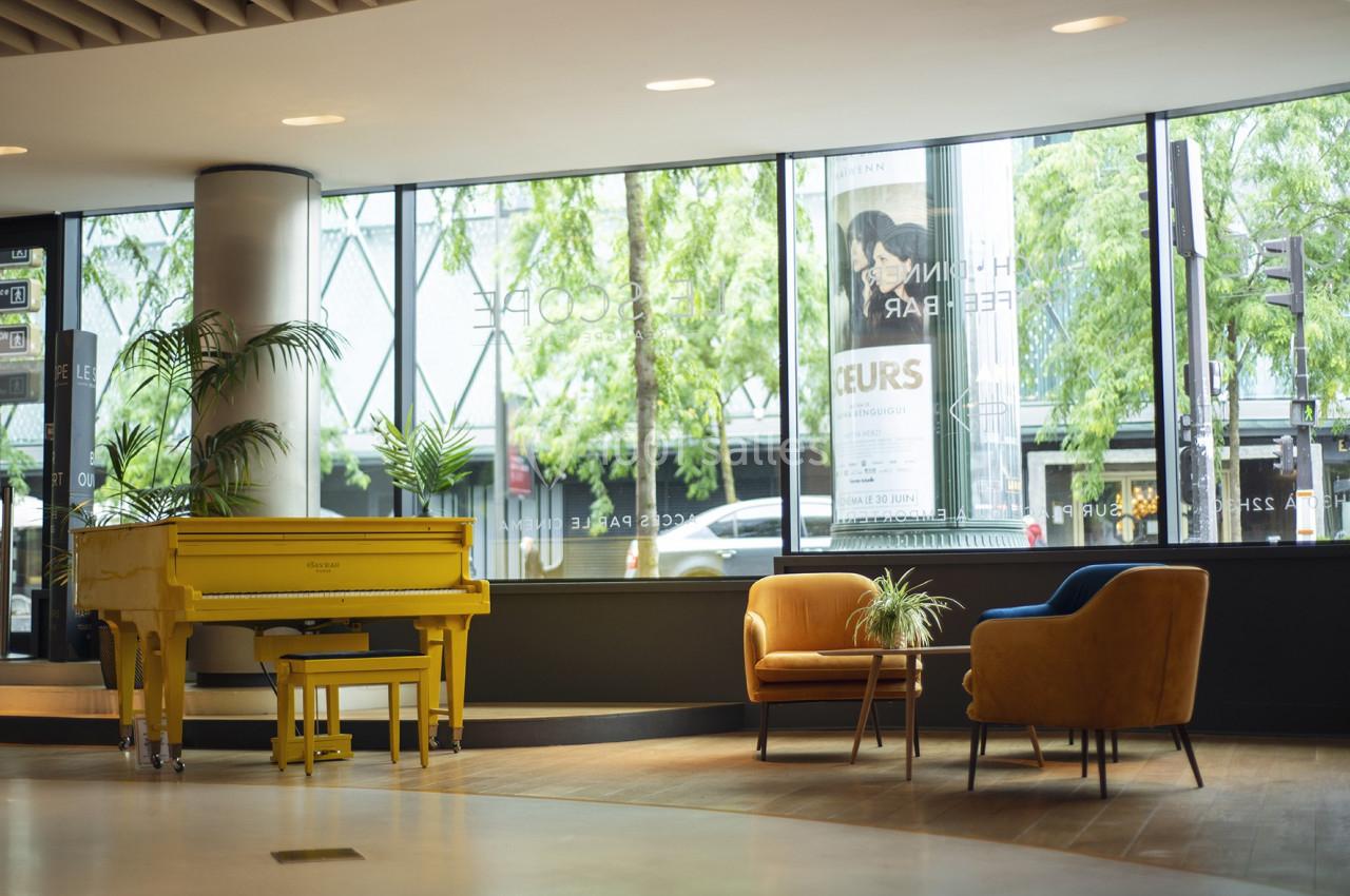 Espace lumineux avec un piano jaune, deux fauteuils et une table basse, entourés de grandes baies vitrées donnant sur des…