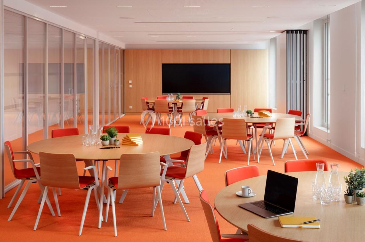 Salle de réunion lumineuse avec tables rondes, chaises orange, écran noir et éléments de décoration modernes.